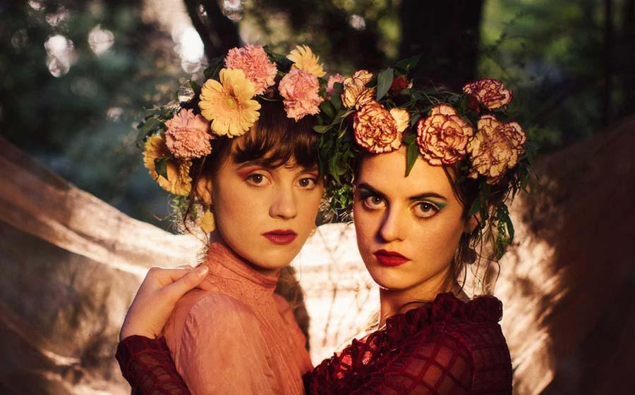 two-women-standing-in-front-of-each-other-wearing-crowns-made-of-flowers-with-their-heads-turned-toward-the-camera