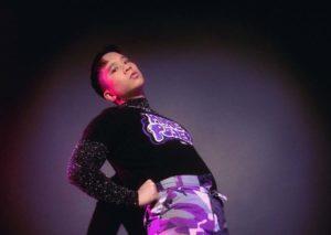 Man fdressed in black and purple standing in a dark room with his hands on his hips