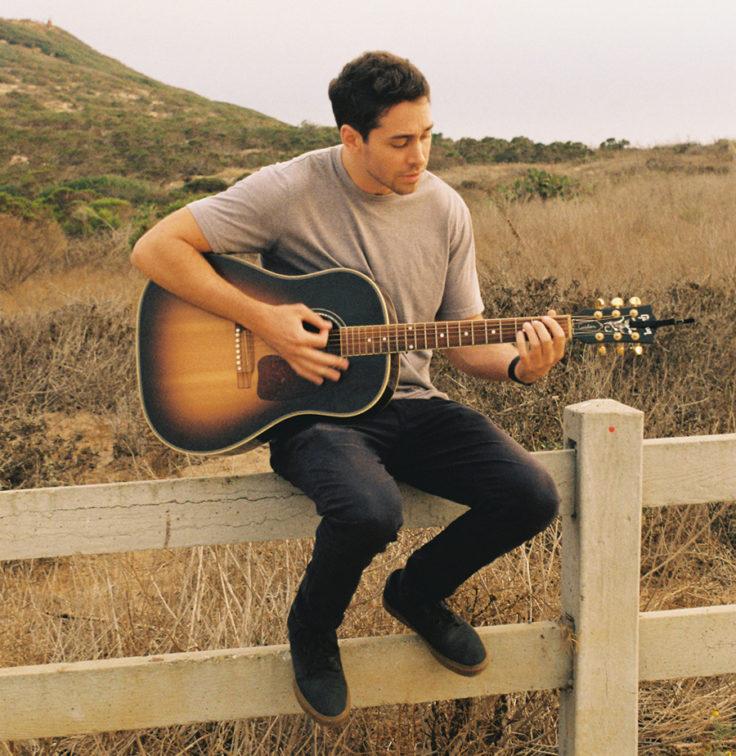 man sitting on a fence in the countryside playing guitar