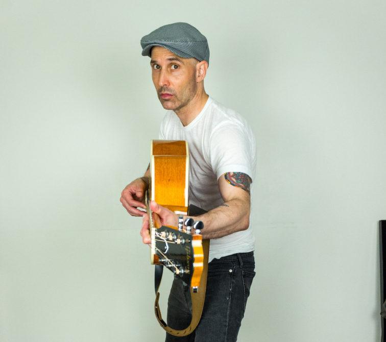 man standing in front of a creamy white wall pointing his guitar neck at the camera