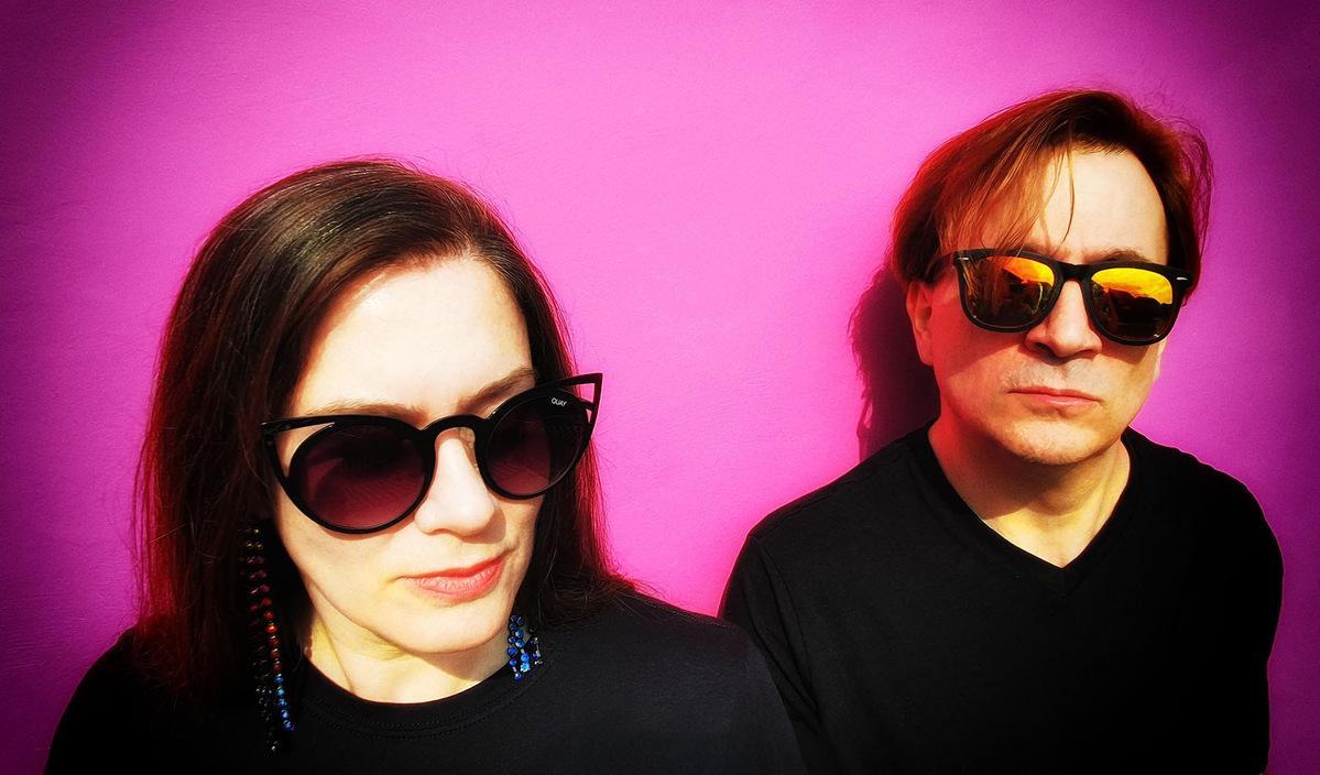 headshot of man and woman standin gin front of a blanck purple wall