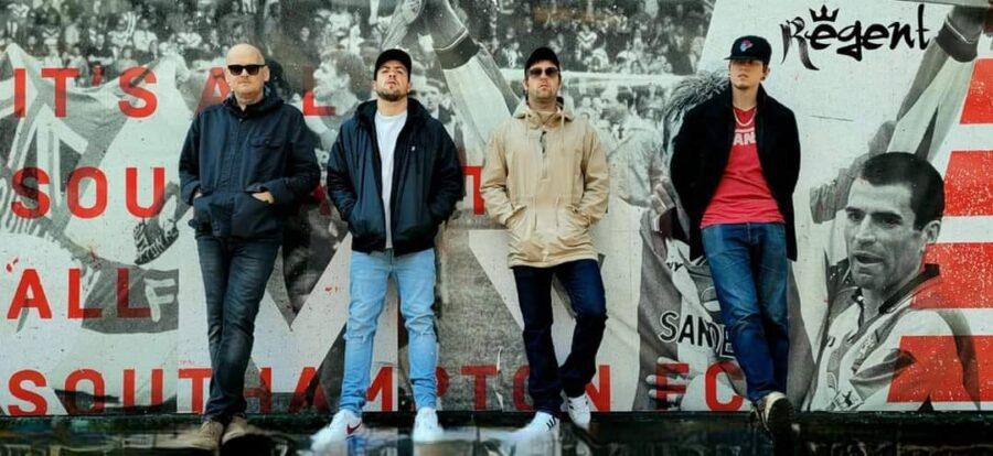 4 male band members standing in front of a cement wall on the street