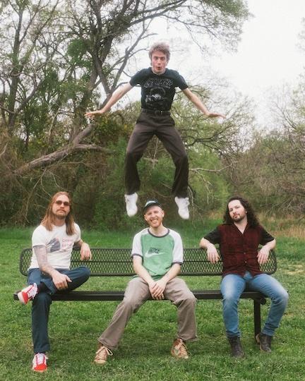 man jumping behind 3 men sitting on a bench in a park