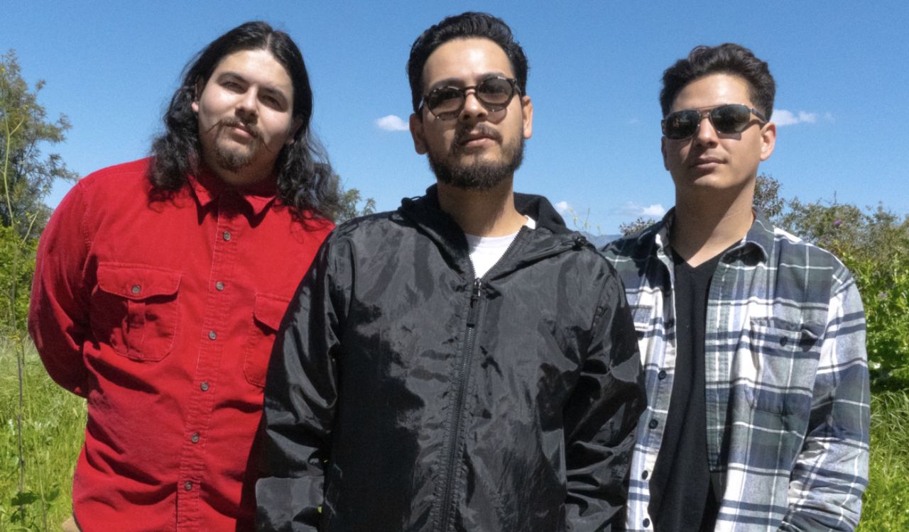 three band members standing outside on a sunny day in front of trees